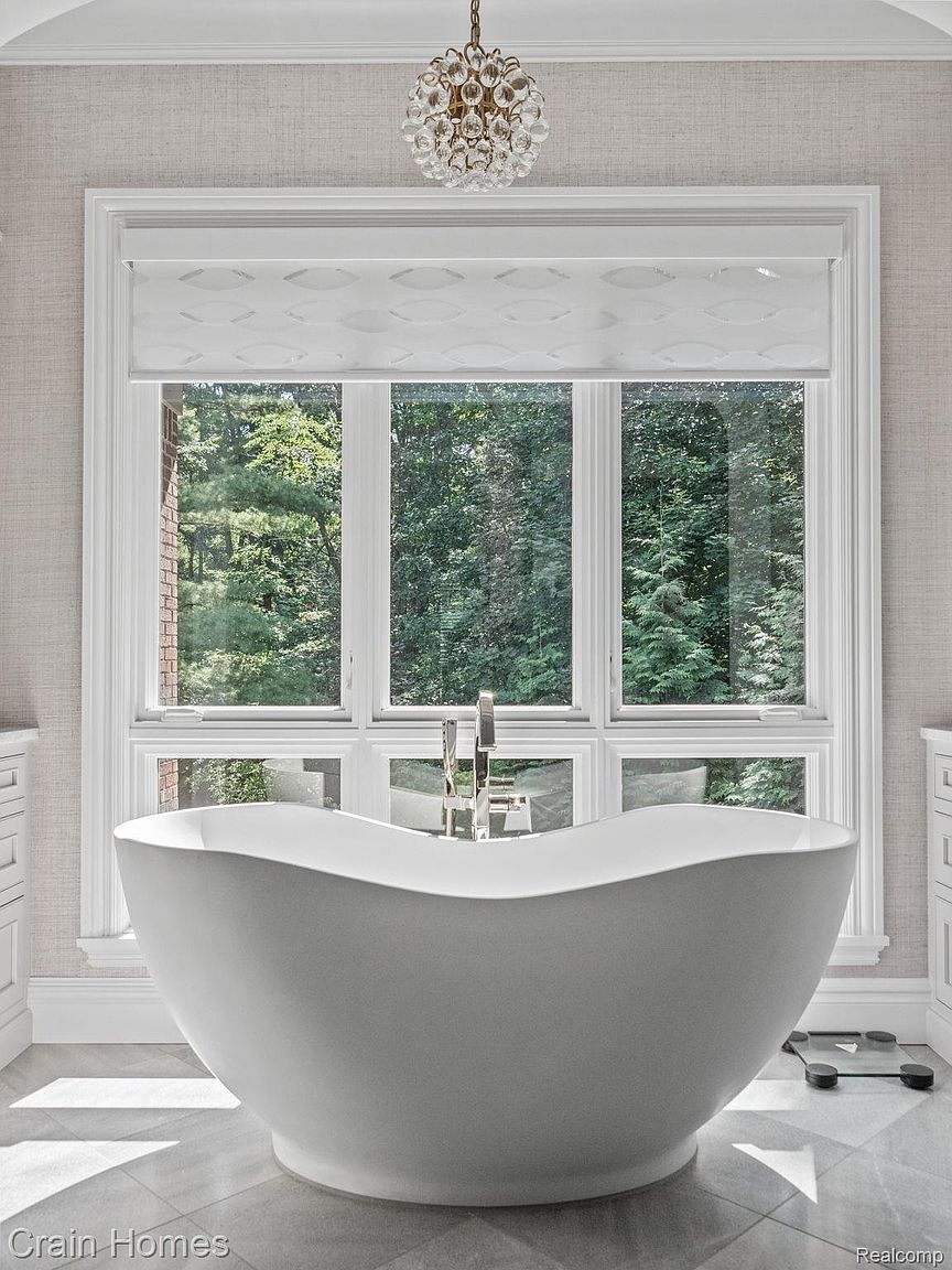 This is a luxurious primary bathroom featuring a freestanding white bathtub positioned in front of a large window with a view of lush greenery. The room is decorated in neutral tones with a textured wallpaper, and a modern chandelier hangs from the ceiling. The overall impression is one of tranquility and spa-like elegance.