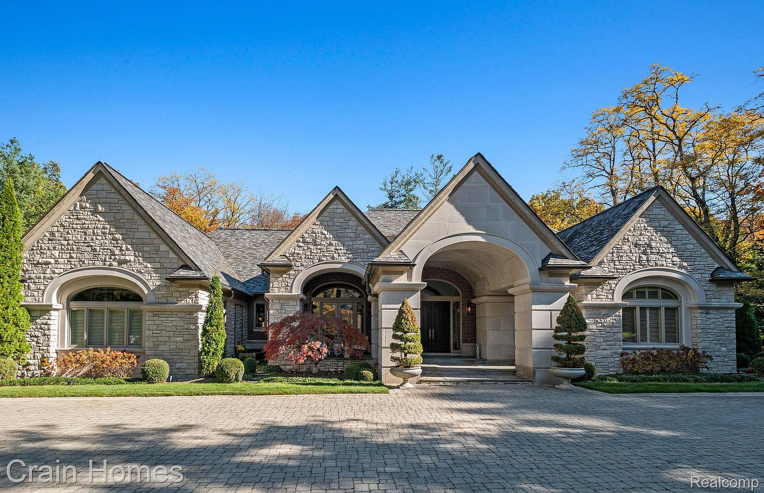 This is a front exterior view of a luxurious stone house with a prominent arched entryway. The house features multiple gabled roofs, arched windows, and meticulously maintained landscaping. The driveway is paved with interlocking stones, adding to the property's curb appeal and upscale aesthetic.