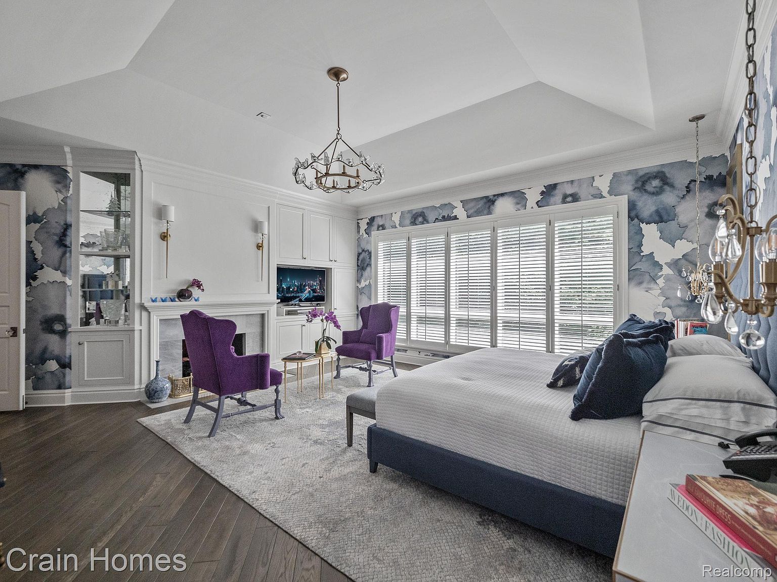 This is a luxurious primary bedroom featuring a king-size bed with a blue base and white quilted bedding, complemented by blue accent pillows. The room is decorated with elegant wallpaper, a crystal chandelier, and two purple armchairs facing a fireplace with a TV above. The space is well-lit with natural light from the shuttered windows and features dark hardwood floors and a light-colored area rug, creating a serene and sophisticated atmosphere.