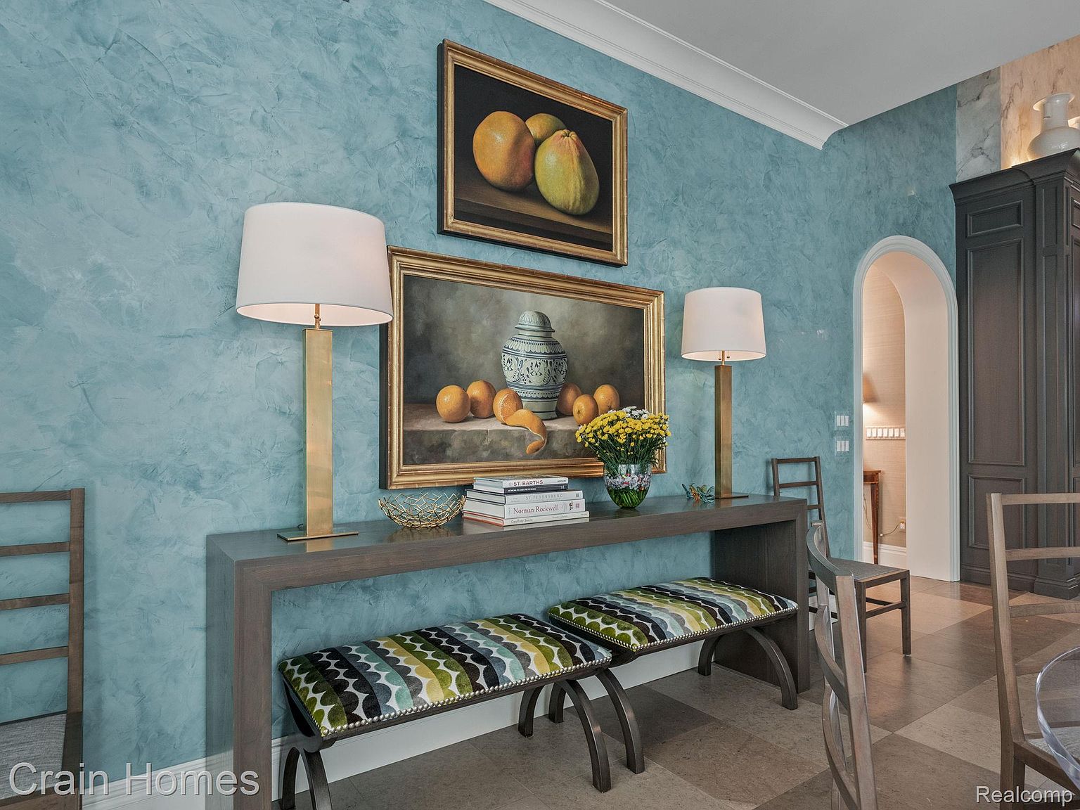 This interior shot showcases a dining room with a sophisticated aesthetic. A console table adorned with lamps and artwork sits against a textured blue wall, complemented by upholstered benches. The room features an arched doorway leading to another space, and a dark wood cabinet adds a touch of elegance.