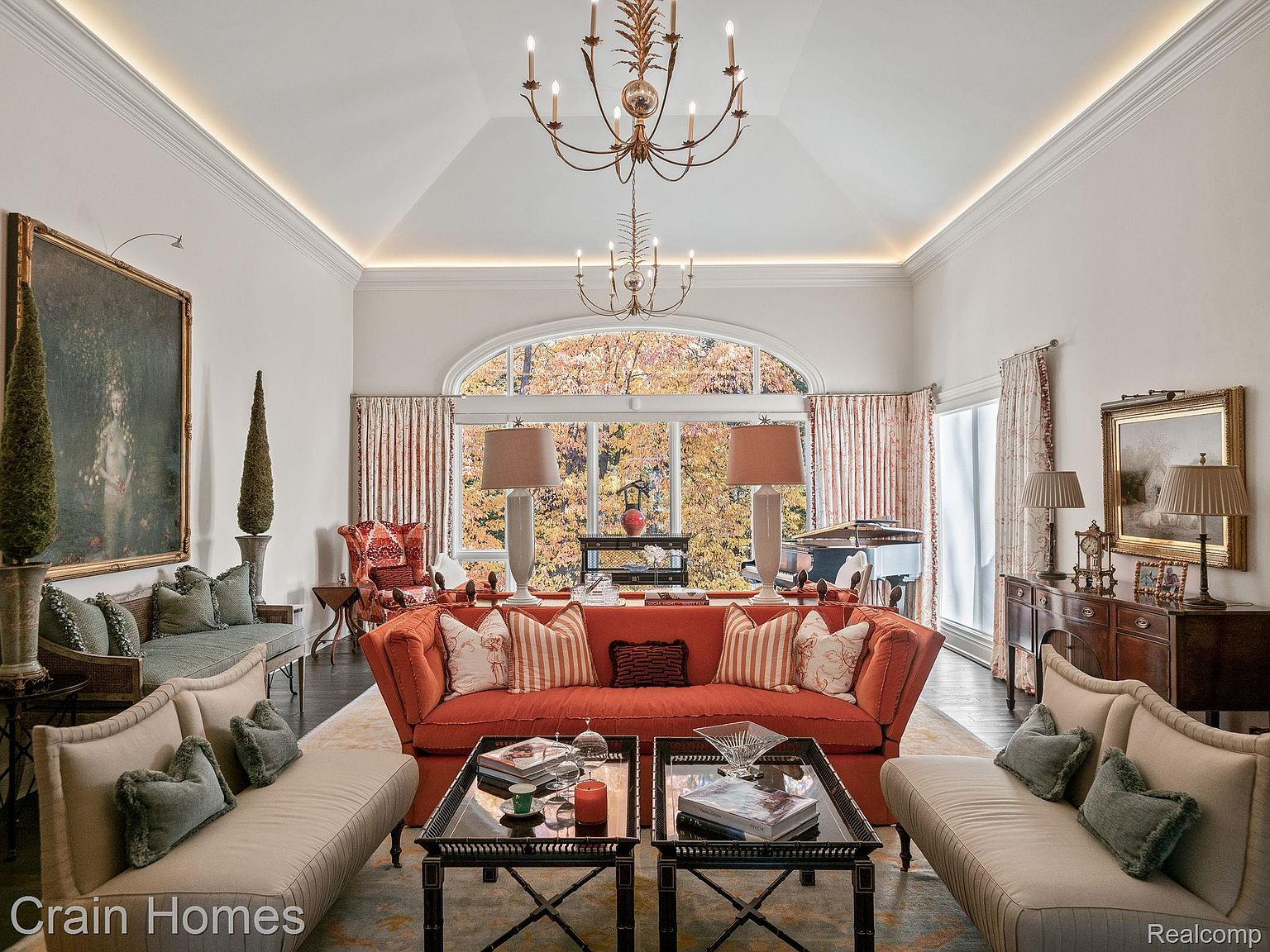This is an interior shot of a luxurious living room featuring an orange sofa as the focal point, flanked by two beige sofas. The room is well-lit with natural light from a large arched window and complemented by elegant chandeliers. The space is decorated with artwork, lamps, and coffee tables, creating a sophisticated and inviting atmosphere.