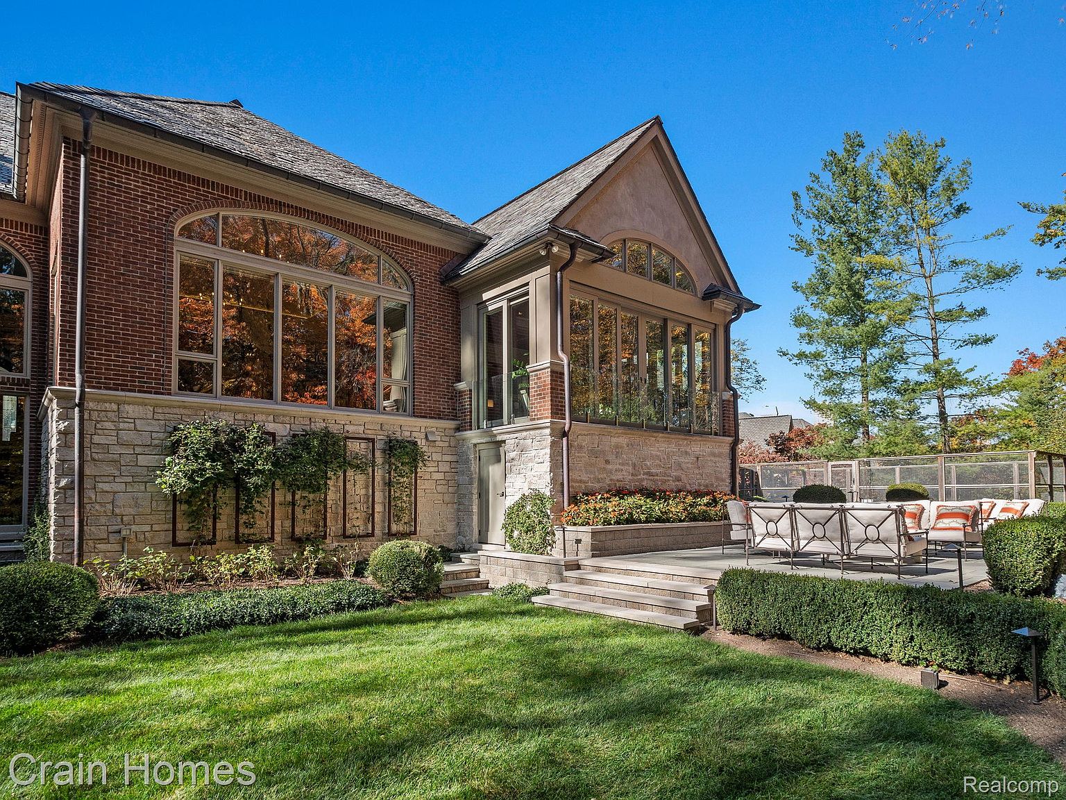 This is a rear exterior view of a luxurious home, showcasing a blend of brick and stone architecture. Large windows reflect the surrounding foliage, and a stone patio with outdoor seating offers a space for relaxation and entertainment. The manicured lawn and landscaping add to the property's appeal.