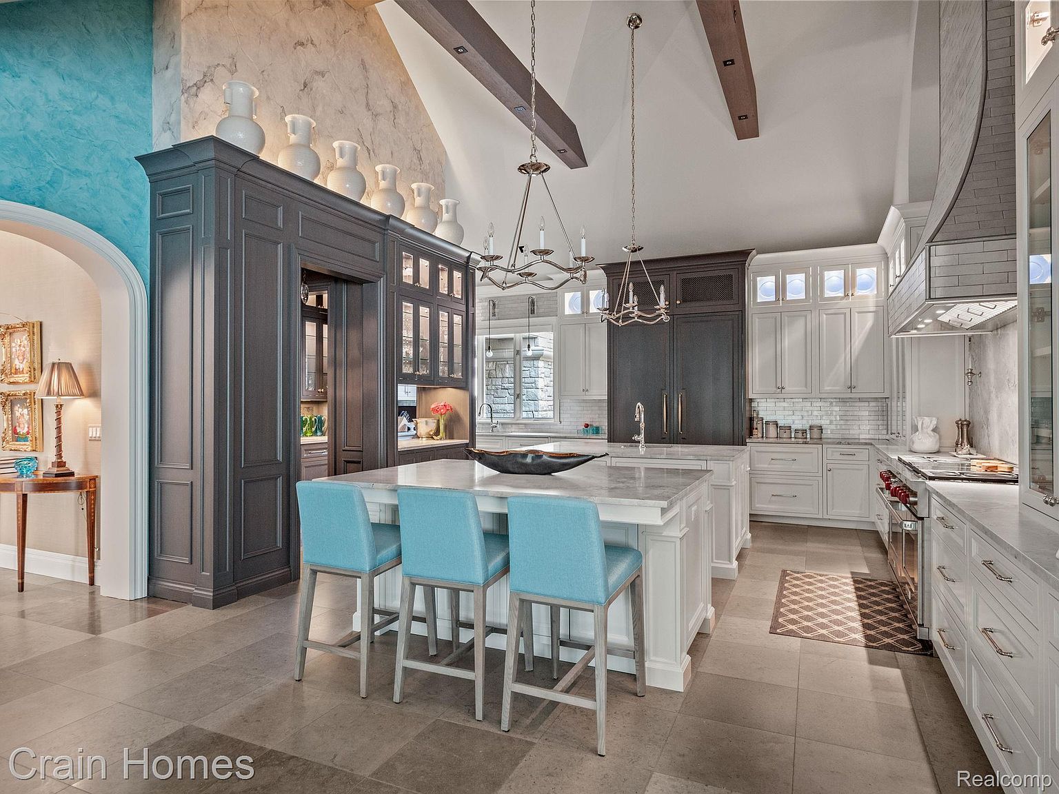 This is a spacious, high-end kitchen featuring a large island with seating, custom cabinetry in both dark gray and white, and marble countertops. The kitchen is well-lit with pendant lighting and recessed lights, and it includes high-end appliances. The overall impression is one of luxury and sophistication.