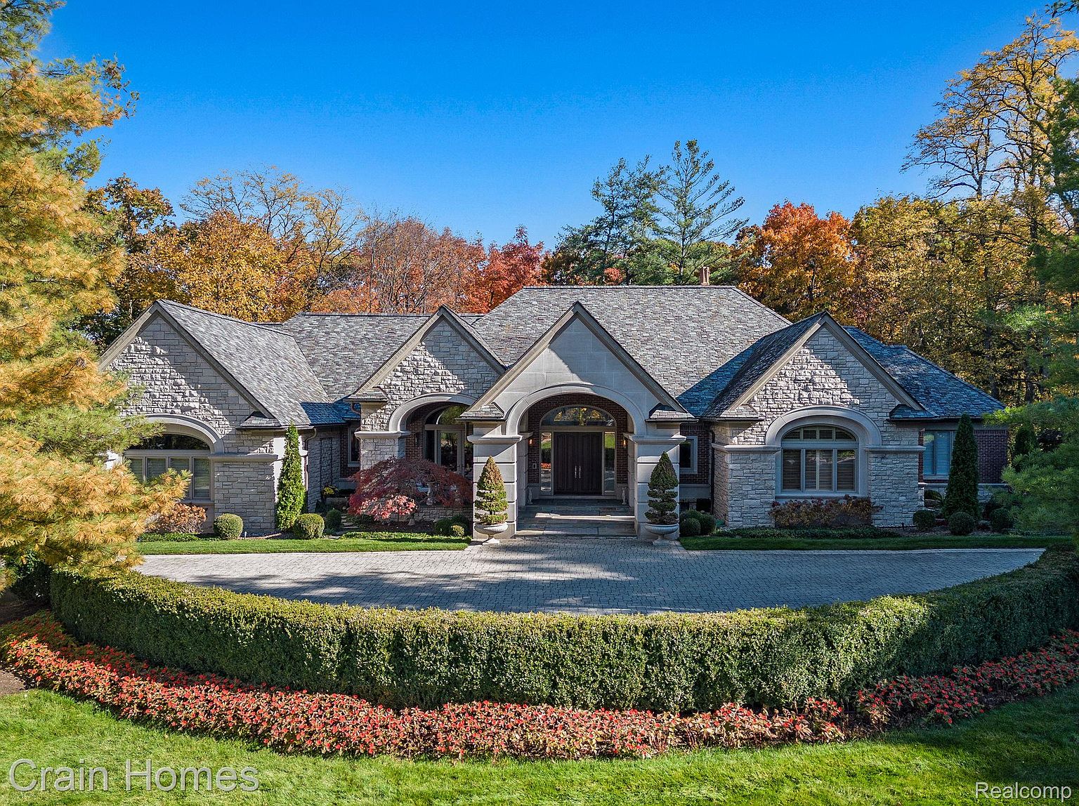 This is a front exterior view of a luxurious stone house with a well-manicured circular driveway and landscaping. The house features a complex roofline with multiple gables, arched windows, and a grand entryway. The surrounding trees display vibrant fall colors, adding to the property's curb appeal.