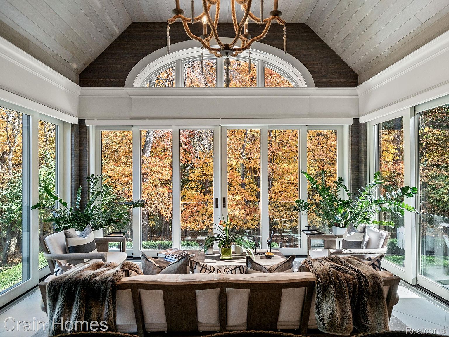 This is a bright and airy living room featuring large windows that offer a stunning view of autumn foliage. The room is furnished with comfortable seating, including a sofa adorned with a fur throw, and is decorated with plants, creating a serene and inviting atmosphere. A unique chandelier hangs from the vaulted ceiling, adding a touch of rustic elegance.