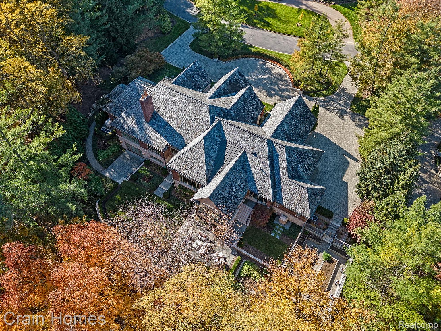 This aerial view showcases a sprawling residence with a complex, multi-gabled roof design and a brick exterior. The property is surrounded by mature trees displaying vibrant fall colors, and a winding driveway leads to the house. The meticulously landscaped grounds feature manicured gardens and stone pathways, enhancing the estate's curb appeal.