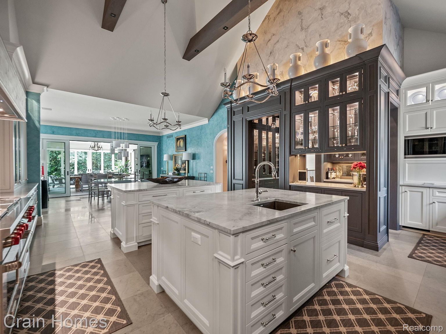 This is a spacious, high-end kitchen featuring two large islands with marble countertops and white cabinetry. A dark wood cabinet with glass doors displays glassware and decorative items, adding a touch of elegance. The kitchen opens to a dining area with a view of the outdoors, creating a seamless transition between spaces.