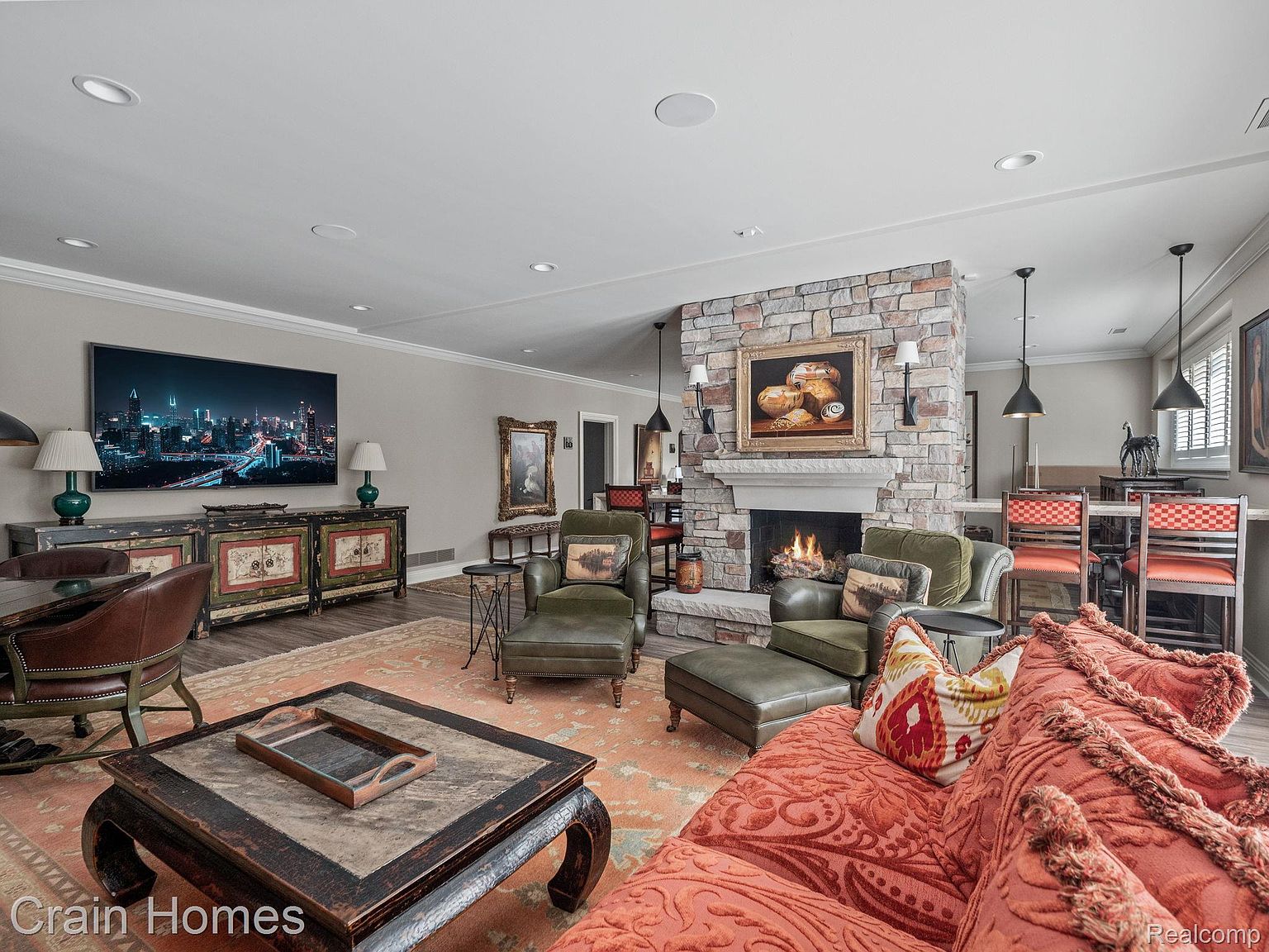 This is an interior shot of a living room featuring a stone fireplace with a painting above it, flanked by two green armchairs and a matching ottoman. A large, ornate coffee table sits in the foreground, and a comfortable-looking sofa with decorative pillows is visible on the right. A TV is mounted on the wall above a cabinet, and the room is decorated with lamps and artwork, creating a warm and inviting atmosphere.