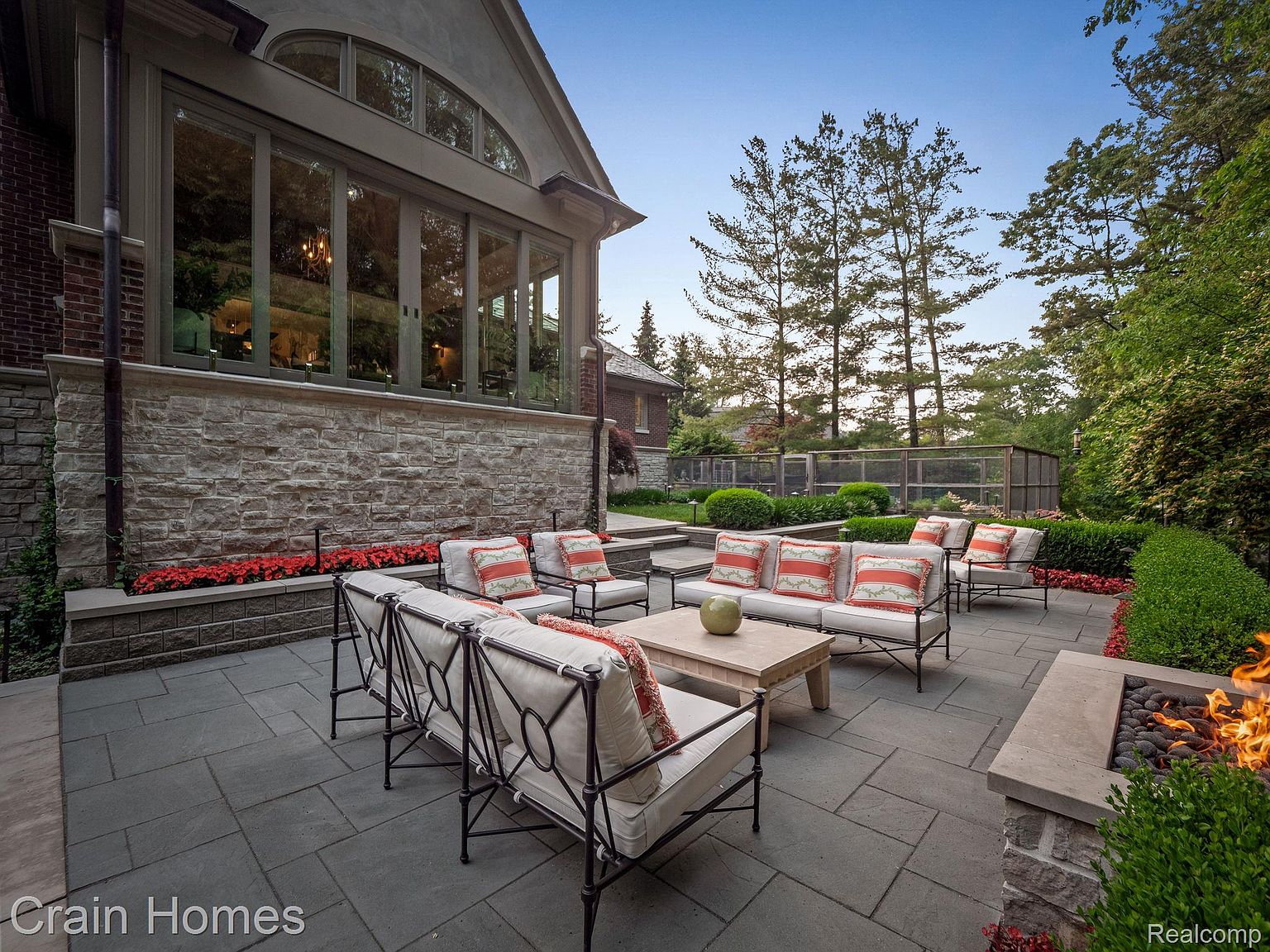 This image showcases a luxurious outdoor patio area, featuring comfortable seating arrangements with plush cushions and decorative pillows. A stone coffee table sits in the center, complemented by a built-in fire pit on the side. The patio is paved with large stone tiles and is surrounded by manicured greenery, creating an inviting and elegant outdoor living space.