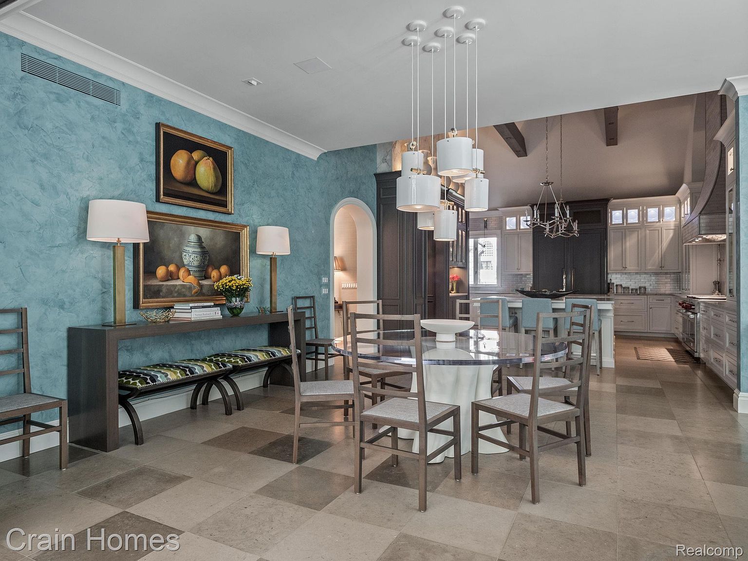 This interior shot showcases a stylish dining room with a round glass-top table surrounded by wooden chairs. A console table with lamps and artwork adorns the wall, complemented by a patterned bench. The room flows seamlessly into a modern kitchen, creating an open and inviting space.