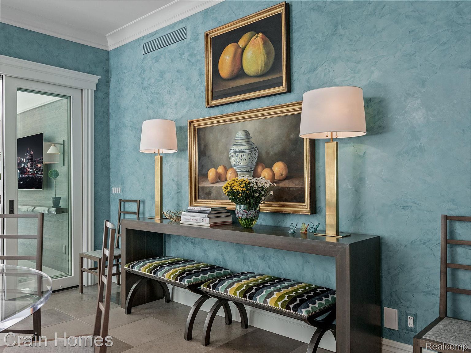 This interior shot showcases a dining area with a sophisticated aesthetic. A console table adorned with lamps, artwork, and decorative items sits against a textured blue wall, complemented by two upholstered benches. The room features a blend of classic and contemporary elements, creating an inviting and stylish space.