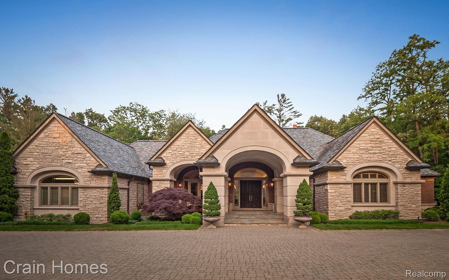 This is a front view of a luxurious stone house with a symmetrical design. The house features multiple gabled roofs, arched entryways, and meticulously manicured landscaping. The driveway is paved with interlocking bricks, adding to the property's curb appeal and creating a grand entrance.