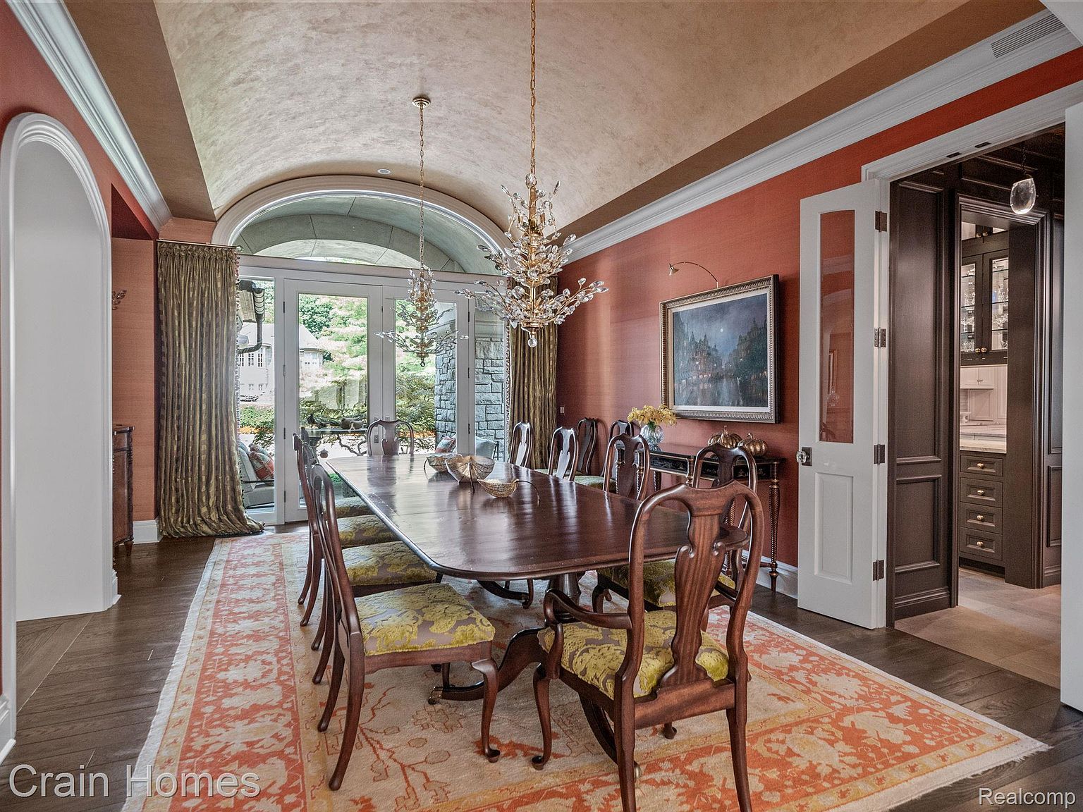 This is an elegant dining room featuring a long, dark wood table set for a formal meal, surrounded by ornate wooden chairs with patterned cushions. Two crystal chandeliers hang above the table, and a large painting adorns the rust-colored walls. The room opens to an outdoor area through arched glass doors, and a doorway leads to another room, showcasing the home's luxurious and spacious design.