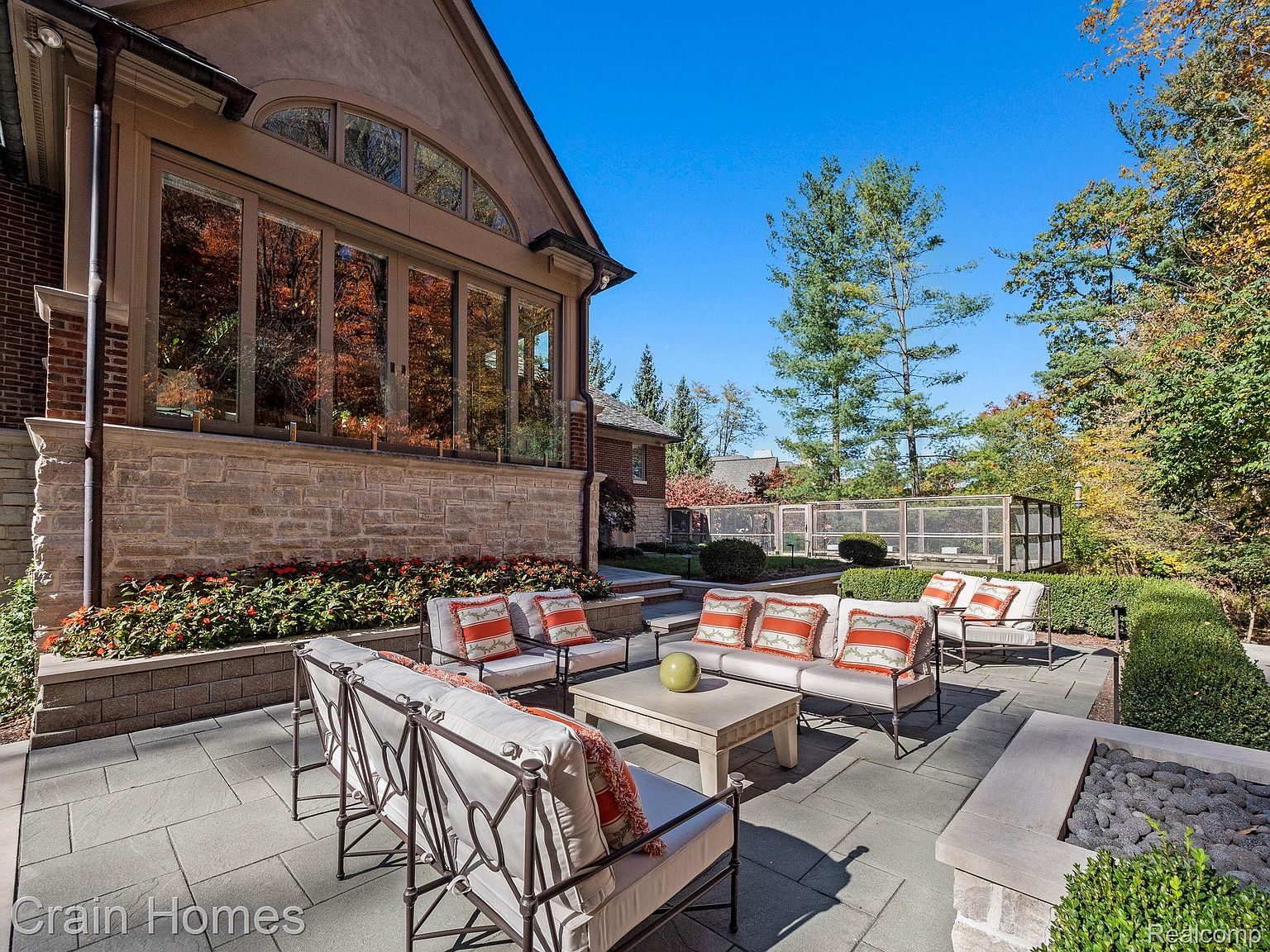 This image showcases a beautifully designed outdoor patio area. The patio features comfortable seating arrangements with plush cushions and decorative pillows, centered around a stone coffee table. The space is surrounded by manicured landscaping, including flower beds and trimmed hedges, creating a serene and inviting atmosphere perfect for relaxation and entertaining.
