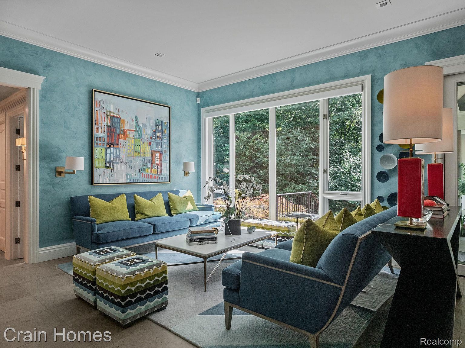 This is an interior shot of a living room featuring a blue sofa with yellow pillows, a modern coffee table, and a large window offering a view of lush greenery. The walls are painted in a textured blue, complemented by white trim and an abstract painting, creating a serene and stylish atmosphere. The room is well-lit and appears spacious, making it an inviting area for relaxation and entertainment.