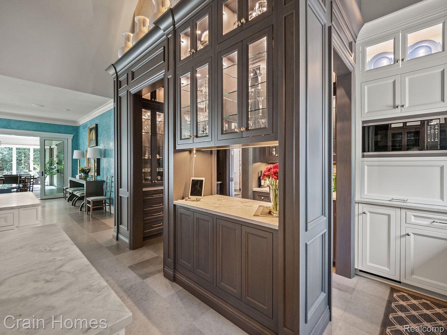This interior shot showcases a sophisticated kitchen area with custom cabinetry in a dark gray finish, complemented by glass-fronted upper cabinets displaying glassware. A light-colored countertop provides a clean contrast, while the overall design exudes elegance and functionality. The perspective gives a view into other rooms, highlighting the open layout.