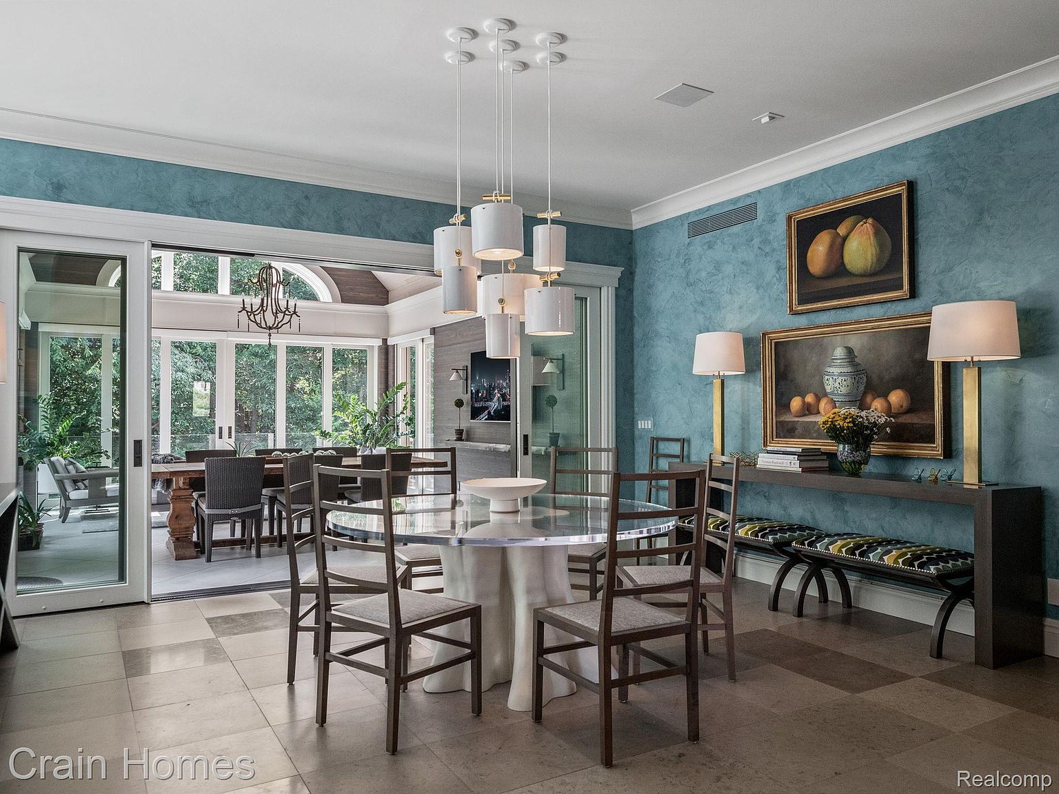 This is an interior shot of a dining room featuring a round glass table with a white tablecloth base, surrounded by wooden chairs with light-colored cushions. A modern chandelier hangs above the table, and a console table with lamps and artwork is positioned against the blue textured wall. The room opens to an adjacent sunroom with a dining set, creating a bright and airy atmosphere.