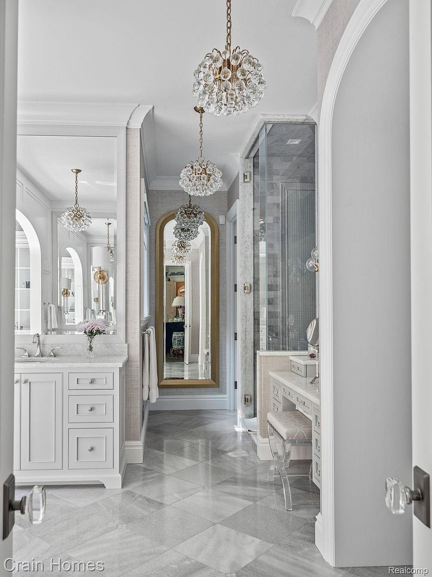 This is a luxurious primary bathroom featuring a vanity with white cabinetry and marble countertops, complemented by a large mirror and elegant crystal chandeliers. The flooring is marble tile laid in a diamond pattern, and a separate vanity area with a clear acrylic stool is visible. The overall impression is one of sophistication and high-end design.