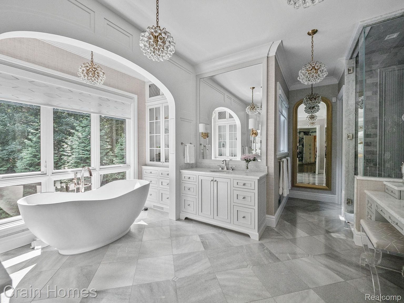 This is a luxurious primary bathroom featuring a freestanding white bathtub positioned in front of a large window offering a view of the outdoors. The bathroom includes a vanity with marble countertops, white cabinetry, and elegant lighting fixtures. A large mirror and an arched doorway leading to another area enhance the spacious and sophisticated feel of the room.