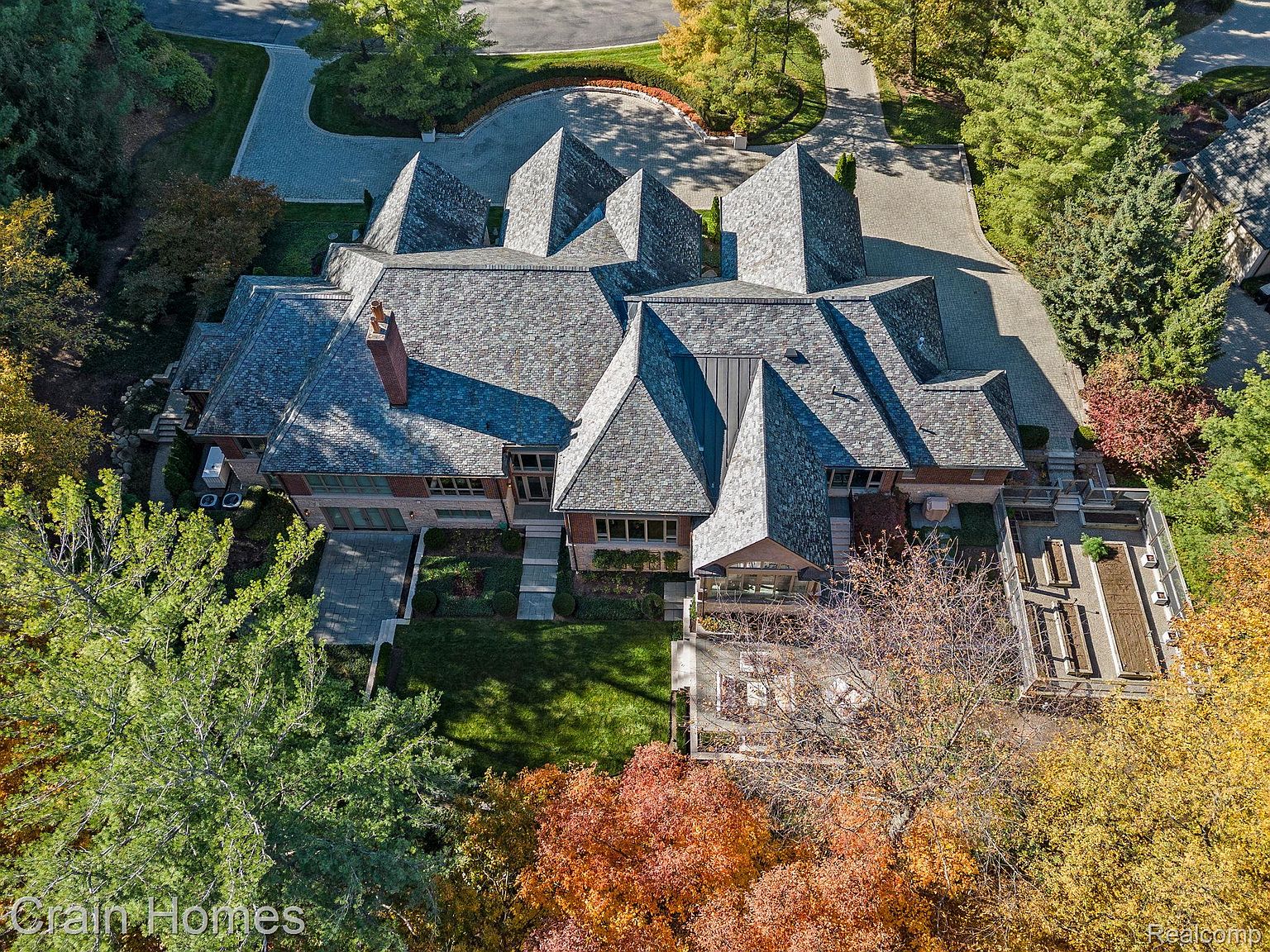 This aerial view showcases a sprawling estate with a complex roofline featuring multiple gables and varying heights. The property includes a well-manicured lawn, mature trees displaying fall colors, and a paved driveway leading to the house. A patio area with outdoor furniture and a raised garden bed are visible, adding to the property's appeal.