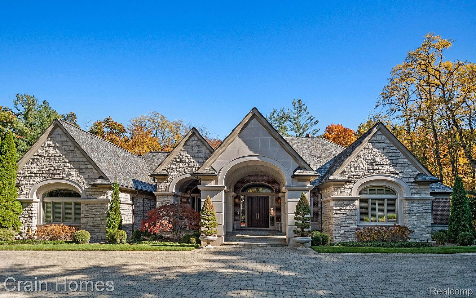 This is a front exterior view of a luxurious stone house with a prominent arched entryway. The house features multiple gabled roofs, arched windows, and meticulously maintained landscaping. The driveway is paved with interlocking stones, adding to the property's curb appeal.