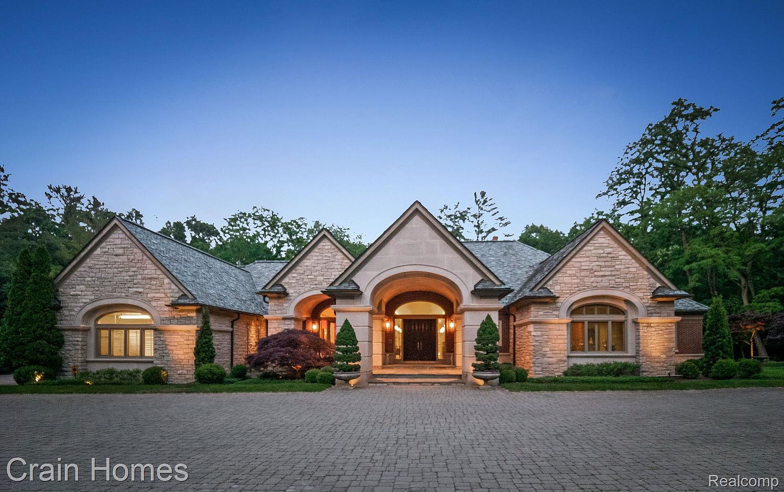 This is a front exterior view of a luxurious stone house at dusk. The house features a prominent arched entryway, multiple gabled roofs, and symmetrically placed windows. The driveway is paved with brick, and the landscaping includes manicured shrubs and trees, creating an inviting and elegant impression.