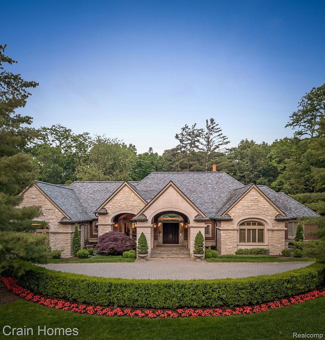 This is a front view of a luxurious single-family home. The house features a stone facade, a prominent arched entryway, and a well-manicured circular driveway with a vibrant flower bed. The landscaping is lush with mature trees, creating a sense of privacy and elegance.
