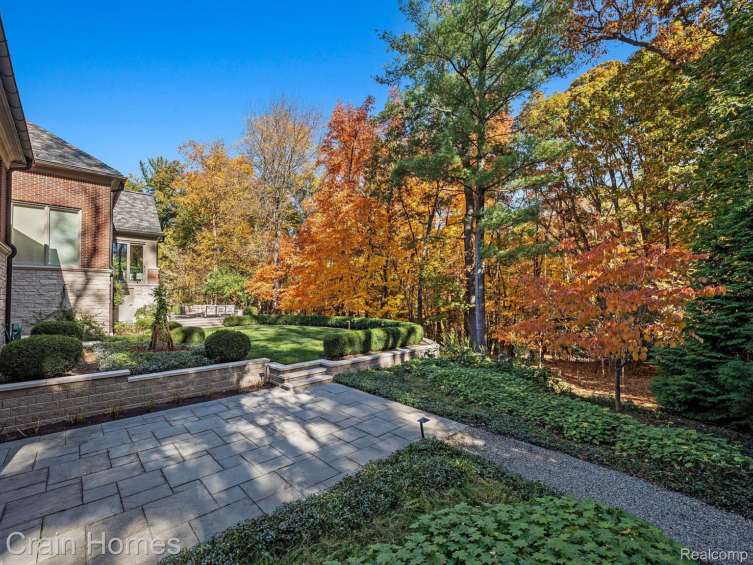 This image showcases a beautifully landscaped backyard featuring a stone patio, manicured lawn, and vibrant fall foliage. The multi-tiered garden beds and mature trees create a serene and private outdoor space. The architectural details of the house blend seamlessly with the natural surroundings, enhancing the property's curb appeal.