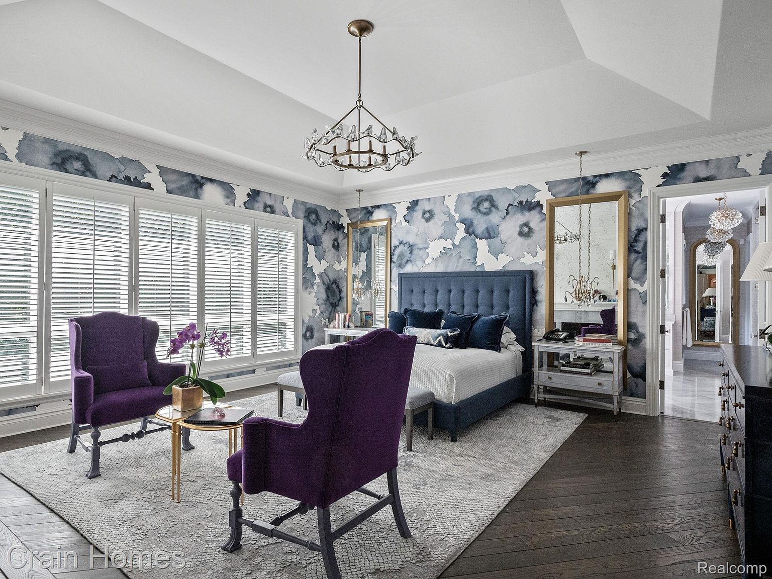 This is a primary bedroom featuring a blue upholstered bed with matching headboard and pillows, flanked by nightstands and mirrors. Two purple armchairs sit near a window with white shutters, and a chandelier hangs from the ceiling. The walls are adorned with a floral wallpaper, and the room is furnished with a patterned rug and dark wood flooring, creating a luxurious and inviting atmosphere.