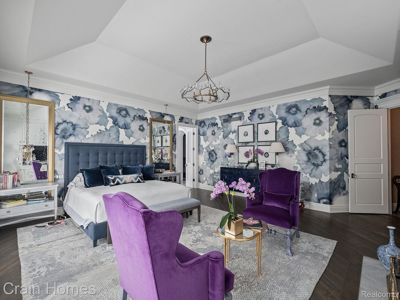 This is a luxurious primary bedroom featuring a king-size bed with a blue upholstered headboard, complemented by a floral wallpaper design. The room is furnished with purple velvet armchairs, a dark blue dresser with framed artwork above, and a crystal chandelier, creating an elegant and sophisticated atmosphere. The perspective is from the foot of the bed, showcasing the room's layout and high-end finishes.