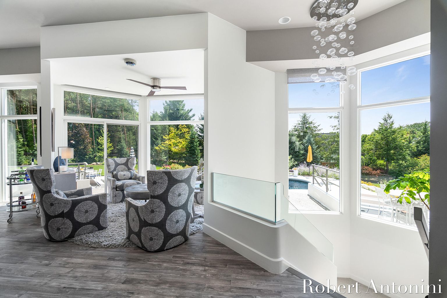 This image showcases a modern living room with a stylish seating area featuring patterned armchairs and a plush rug. Large windows offer abundant natural light and views of the surrounding greenery. The open-concept design flows seamlessly into an adjacent area with a unique chandelier and staircase, creating a bright and inviting atmosphere.