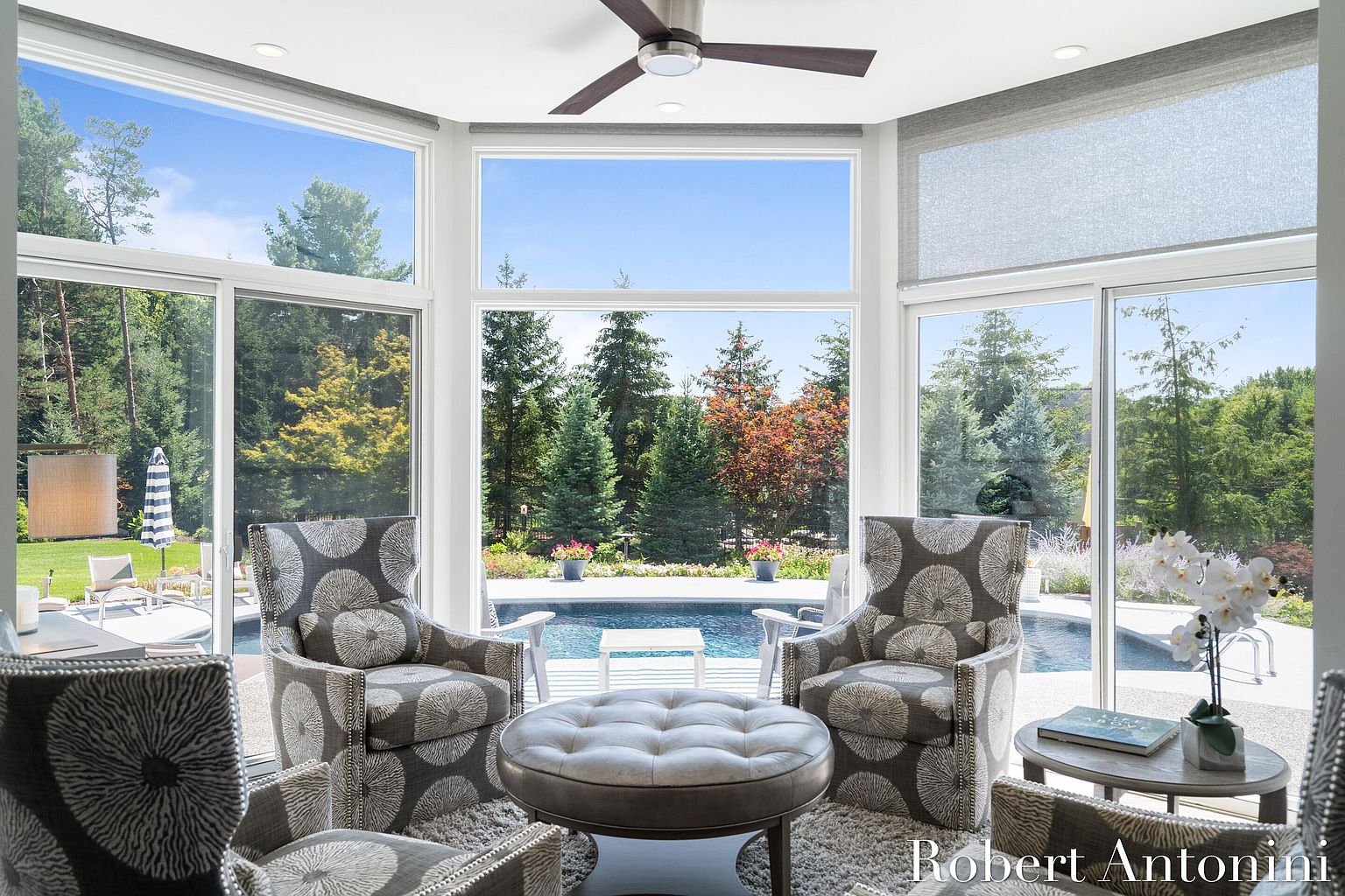This sunroom features large windows offering a view of the pool and surrounding greenery. The room is furnished with patterned armchairs and a round ottoman, creating a cozy seating area. The natural light and outdoor views enhance the appeal of this space, making it a desirable feature for relaxation and entertainment.