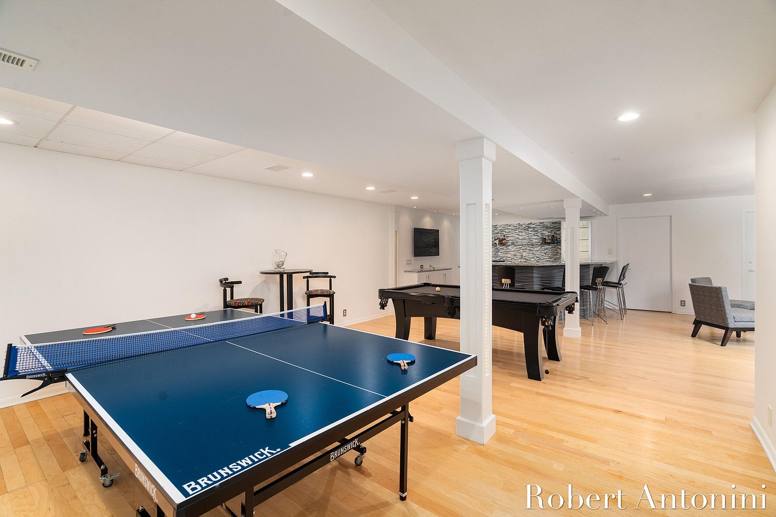 This is a spacious basement featuring a ping pong table and a pool table, suggesting a recreational area. The room has light hardwood floors, white walls, and recessed lighting, creating a bright and inviting atmosphere. A bar area with stools is visible in the background, enhancing the entertainment possibilities of the space.