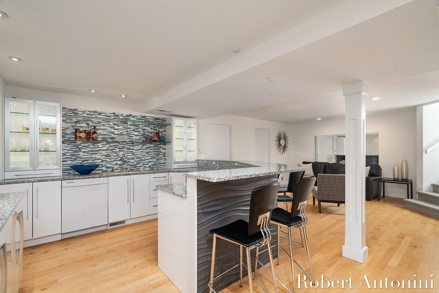This is an interior shot of a finished basement featuring a bar area with white cabinetry, a mosaic tile backsplash, and a granite countertop. The bar has a unique textured front and is complemented by modern bar stools. The space also includes a living area with comfortable seating and hardwood floors, creating a welcoming and stylish entertainment space.