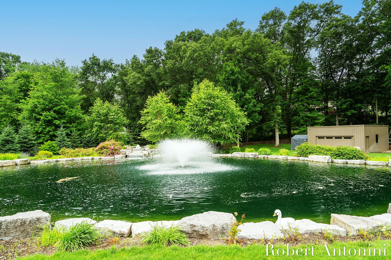 This image showcases a serene yard/garden featuring a large pond with a central fountain, surrounded by lush greenery and mature trees. The pond is lined with natural stones, and a small building is visible in the background, adding to the property's appeal. The overall impression is one of tranquility and natural beauty, making it an attractive feature for potential buyers.