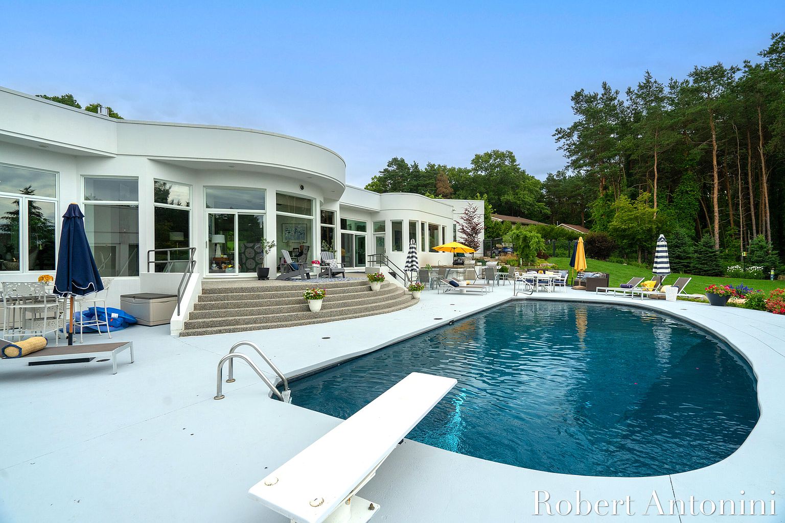 This image showcases a luxurious backyard with a modern, curved white house overlooking a stunning pool. The pool features a diving board and is surrounded by a white deck with various seating arrangements, including lounge chairs and dining sets under umbrellas. Lush greenery and trees provide a serene backdrop, enhancing the property's appeal.