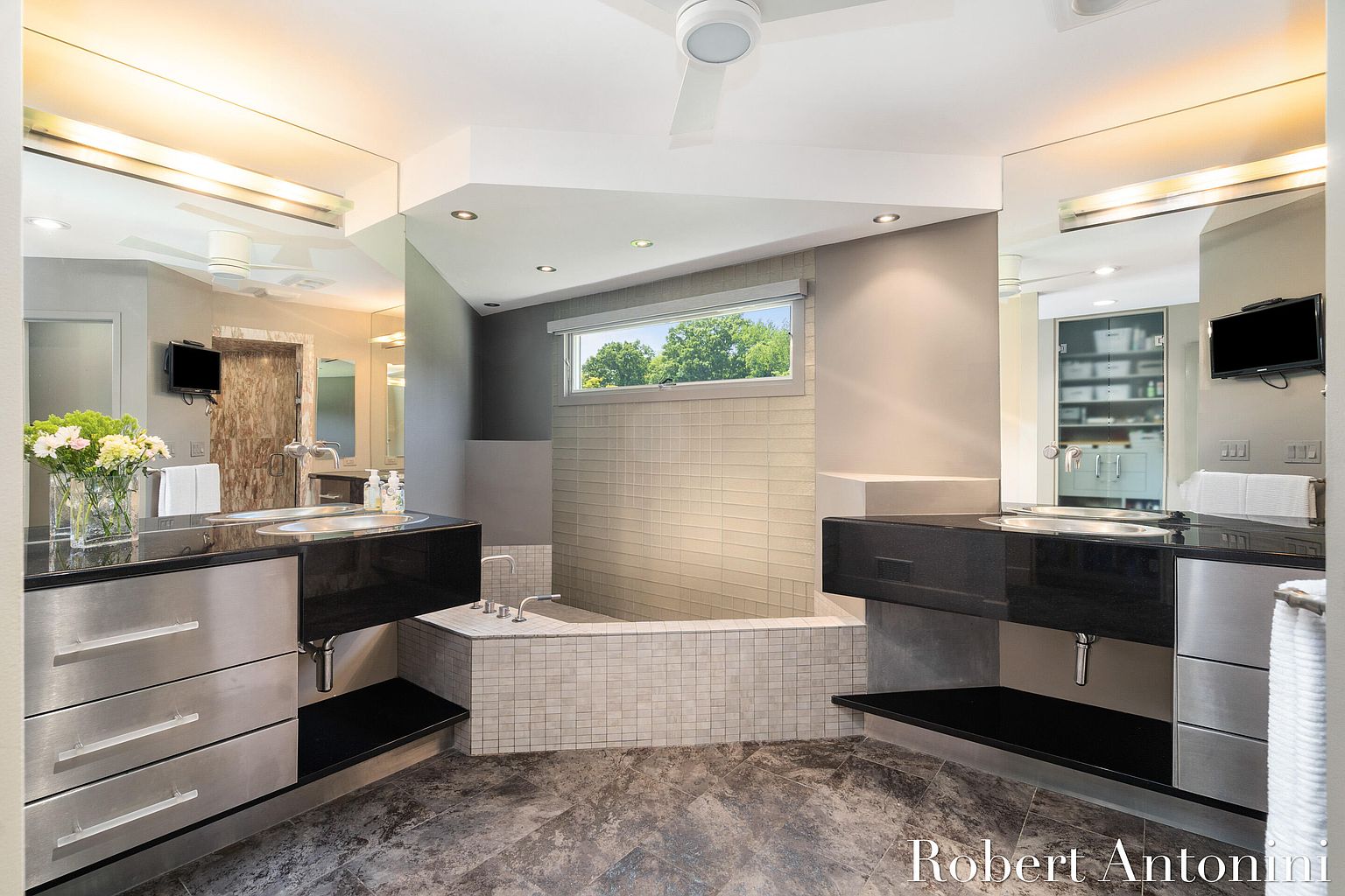 This is a modern primary bathroom featuring dual vanities with black countertops and stainless steel drawers. A large tiled bathtub sits between the vanities, illuminated by a window. The flooring is a dark, patterned tile, and the overall design is sleek and contemporary.