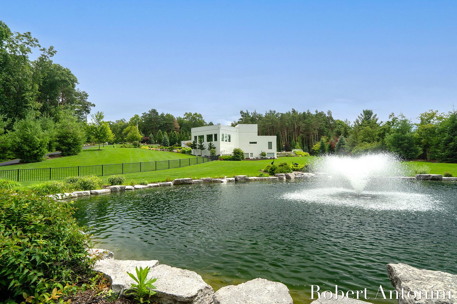 This exterior shot showcases a modern white house set against a lush green lawn and mature trees, creating a serene and private setting. A large pond with a fountain in the foreground adds a luxurious touch, while a black fence provides security and defines the property line. The overall impression is one of tranquility and upscale living.