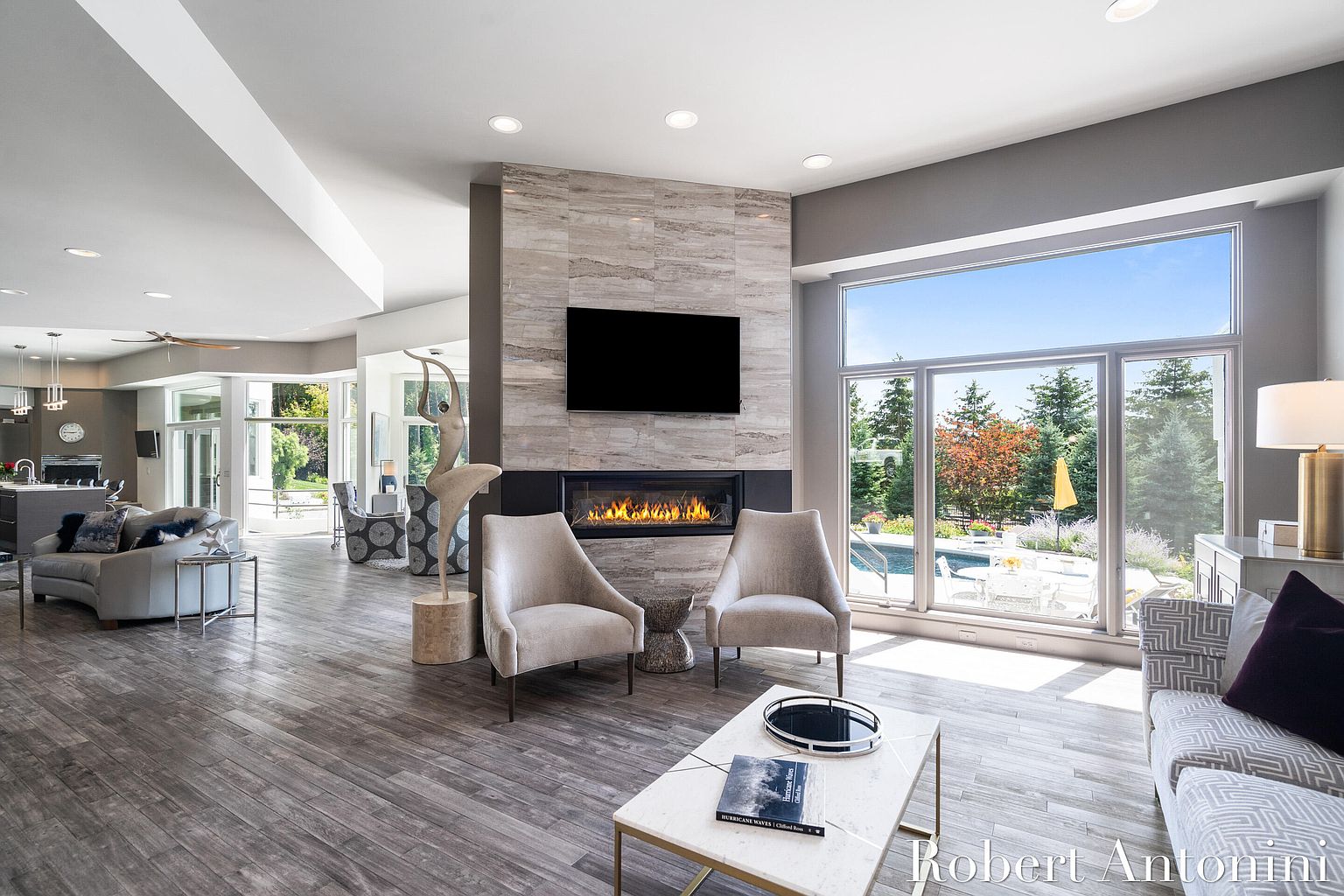 This is an interior shot of a modern living room featuring a fireplace with a mounted television above it. Two armchairs are placed in front of the fireplace, and a coffee table sits in the center of the room. Large windows offer a view of the outdoor pool area, creating a bright and airy atmosphere.