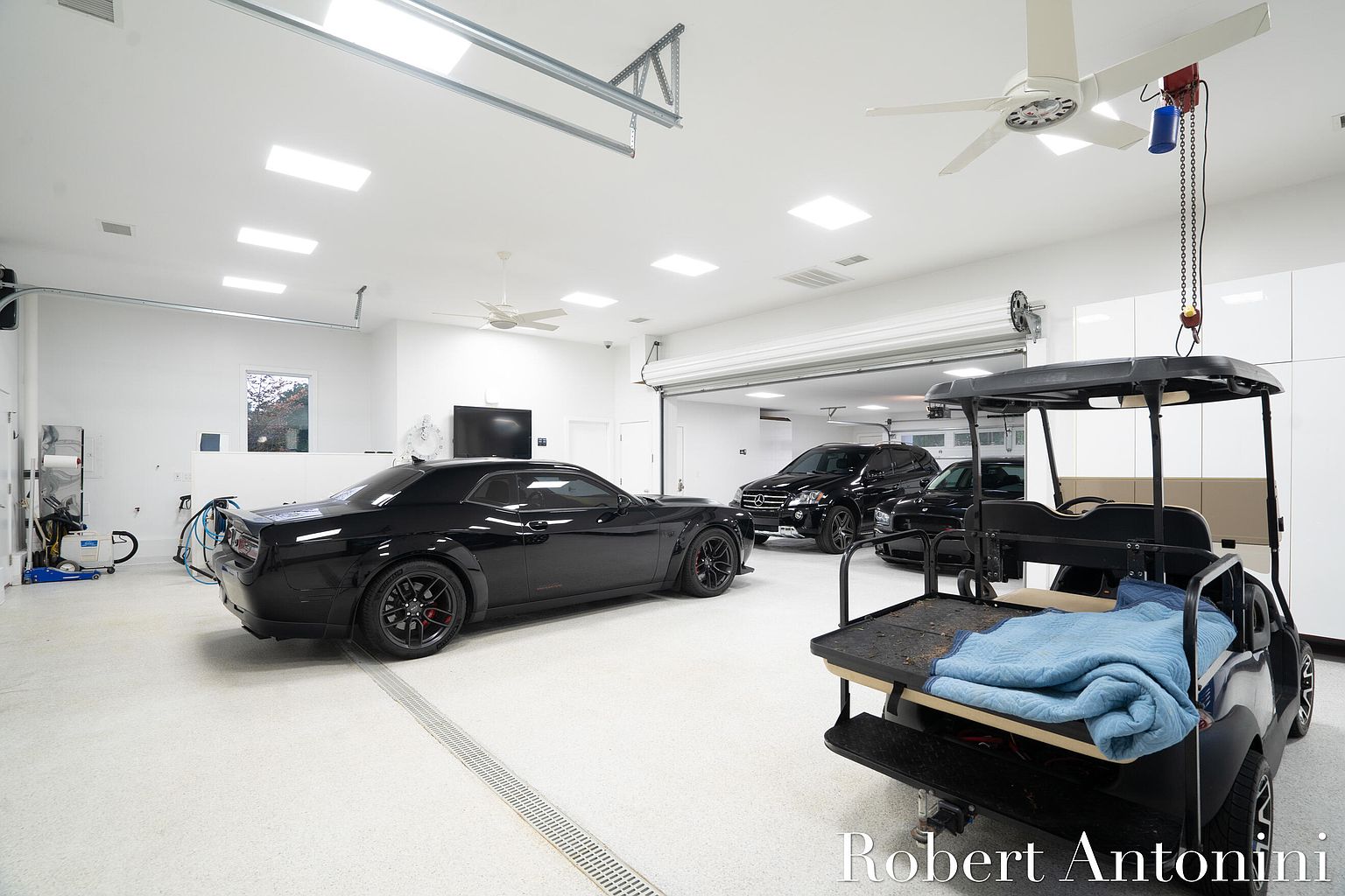 This is a spacious and well-lit garage featuring a black Dodge Challenger, a black Mercedes SUV, and a golf cart. The garage has a clean, modern aesthetic with white walls, a light-colored epoxy floor, and recessed lighting. The space is organized and appears to be climate-controlled, suggesting a high-end property.