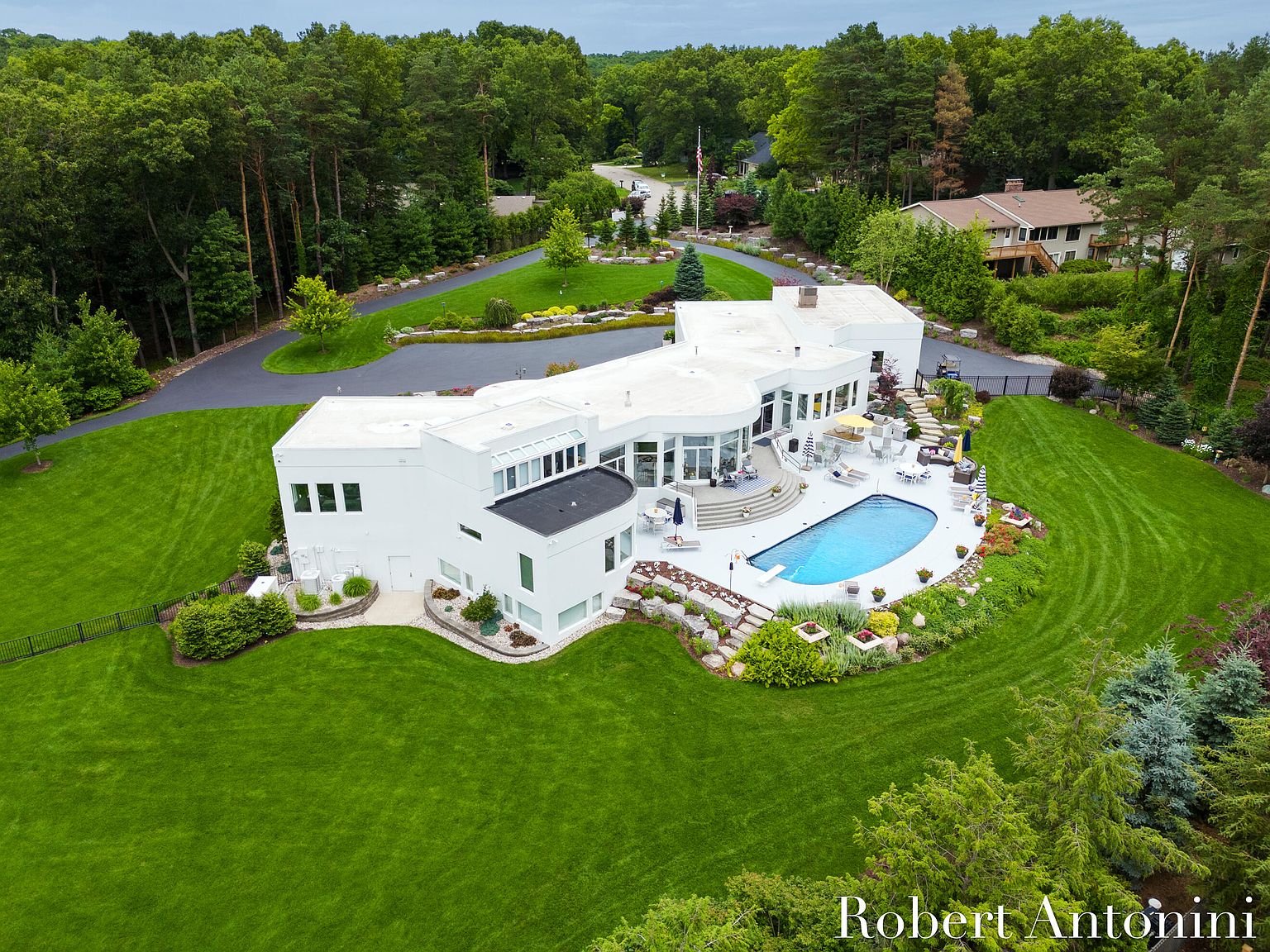 This aerial shot showcases a modern, white mansion with a flat roof, surrounded by lush green lawns and mature trees. A kidney-shaped pool is a focal point in the backyard, complemented by patio furniture and landscaping. A winding driveway leads to the property, enhancing its curb appeal and sense of privacy.