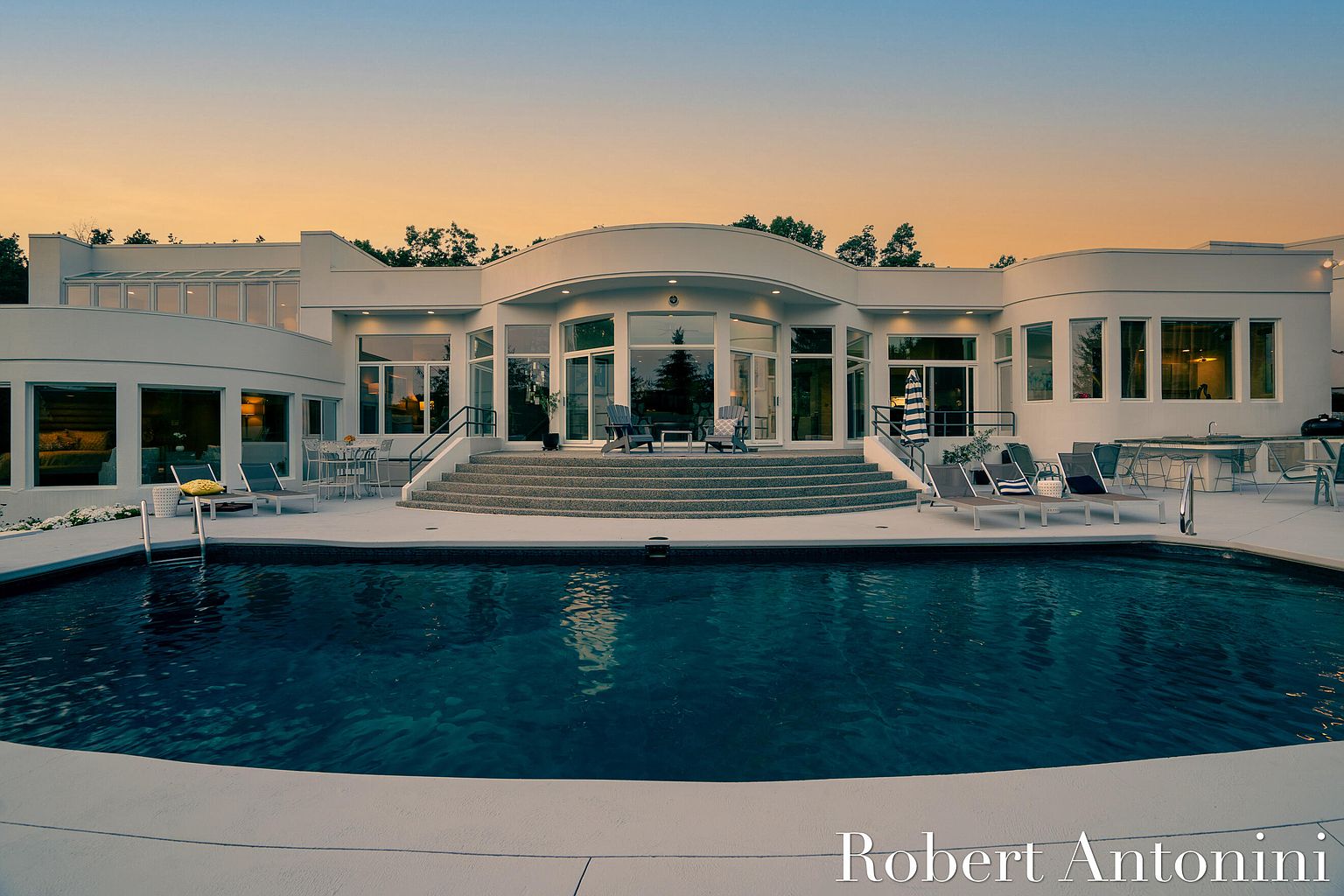 This image showcases the rear exterior of a modern home, emphasizing a luxurious pool area. The pool is surrounded by a spacious patio with lounge chairs and outdoor seating, leading up to a grand entrance with large windows and doors. The architecture is contemporary with curved lines and a clean, white facade, creating an inviting and upscale atmosphere.