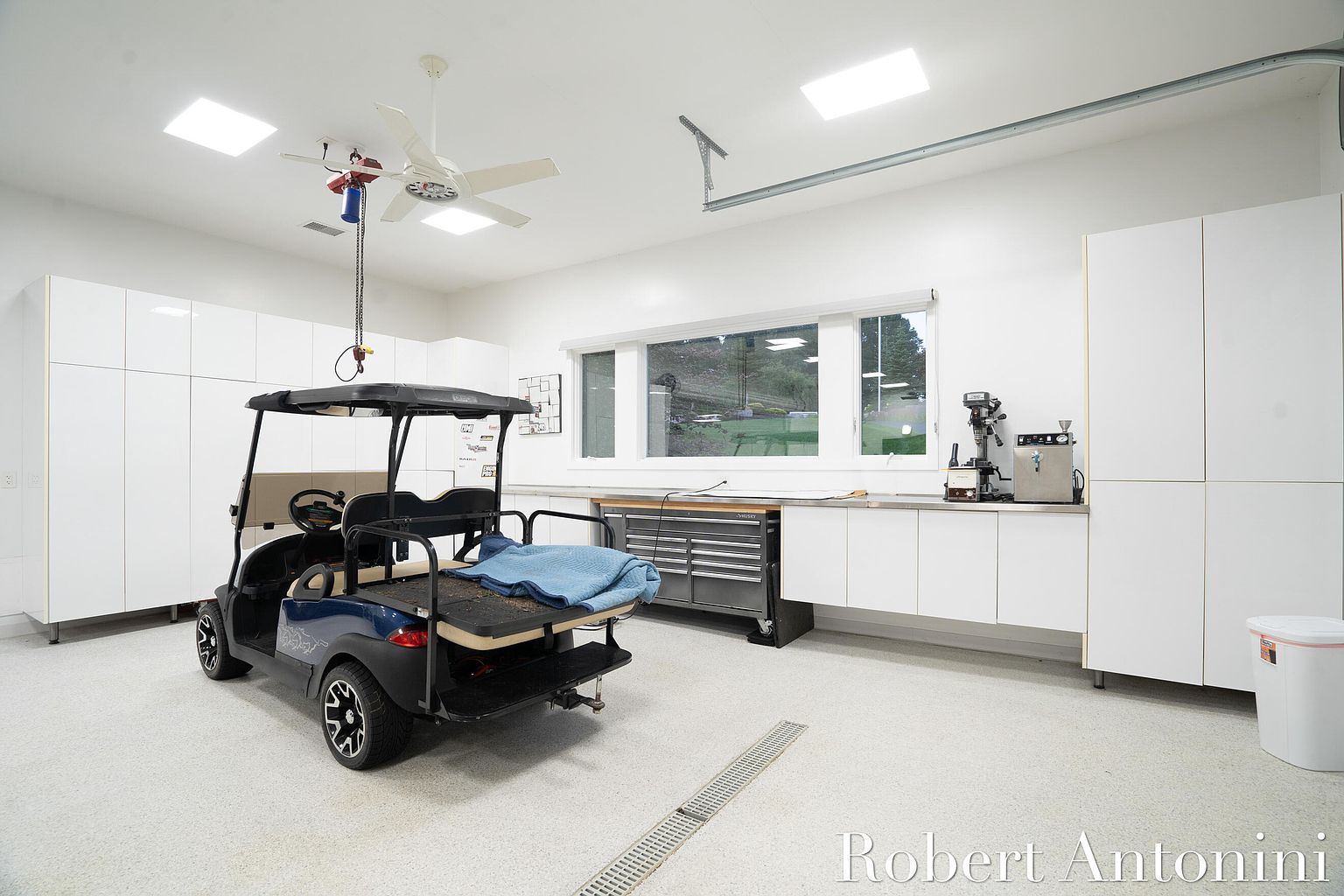 This is a well-organized and brightly lit garage featuring a golf cart parked inside. The garage boasts ample white cabinetry along the walls, providing extensive storage space. A large window offers natural light, and a work area with a stainless steel countertop and tool chest is visible, suggesting a functional and clean workspace.