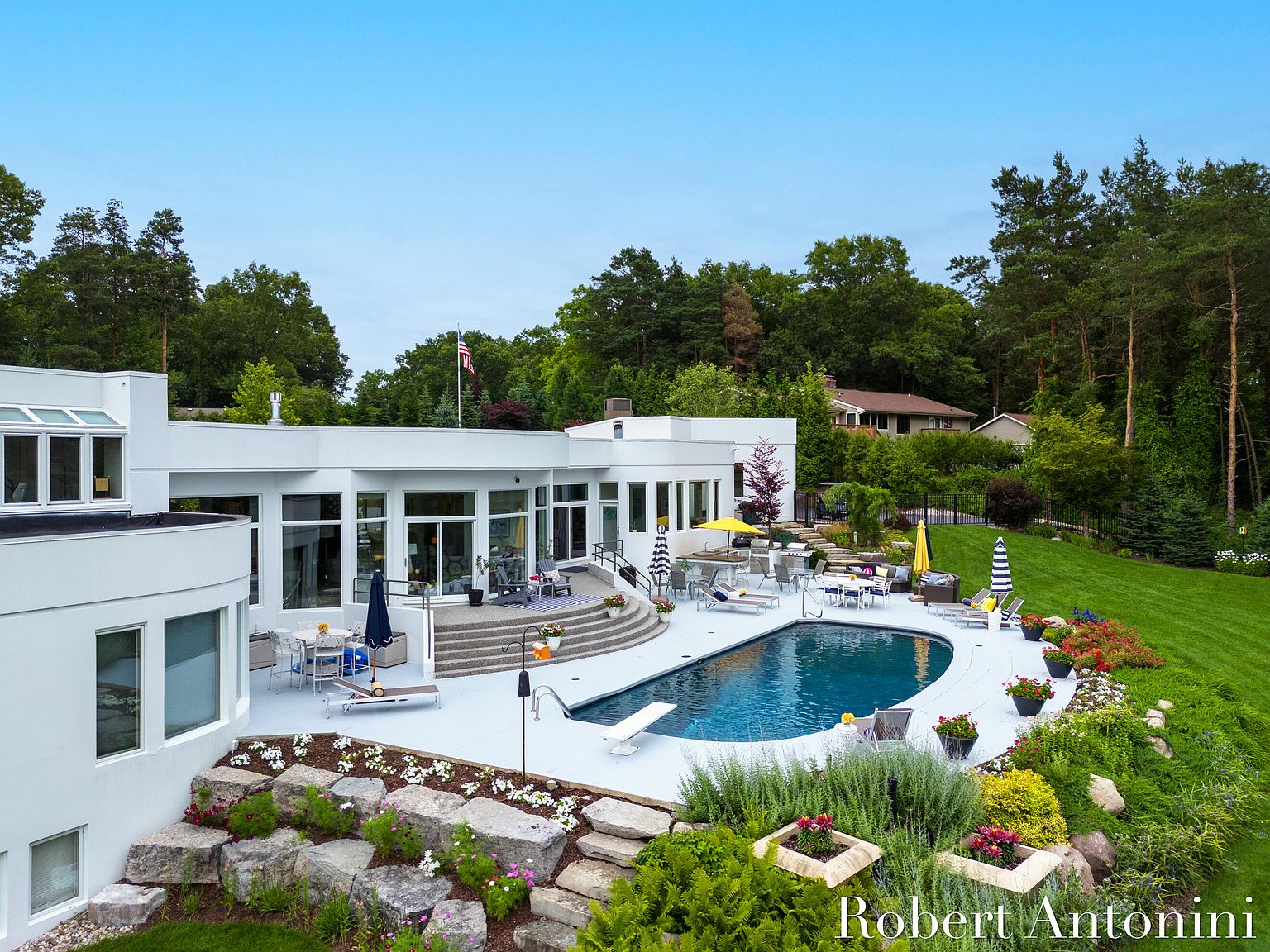 This is an aerial view of a modern home featuring a luxurious outdoor pool area. The pool is surrounded by a white patio with lounge chairs, umbrellas, and dining sets. The landscape includes lush greenery, flower beds, and mature trees, creating a private and serene backyard oasis.