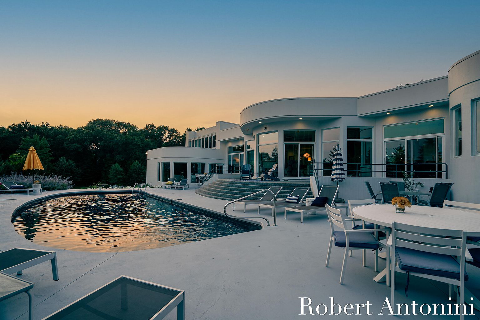 This exterior shot showcases a modern, white-walled home with a curved design, complemented by a luxurious pool and patio area. The pool's dark water reflects the warm hues of the sunset, while lounge chairs and a dining set invite relaxation and entertainment. The image conveys a sense of upscale living and outdoor enjoyment.