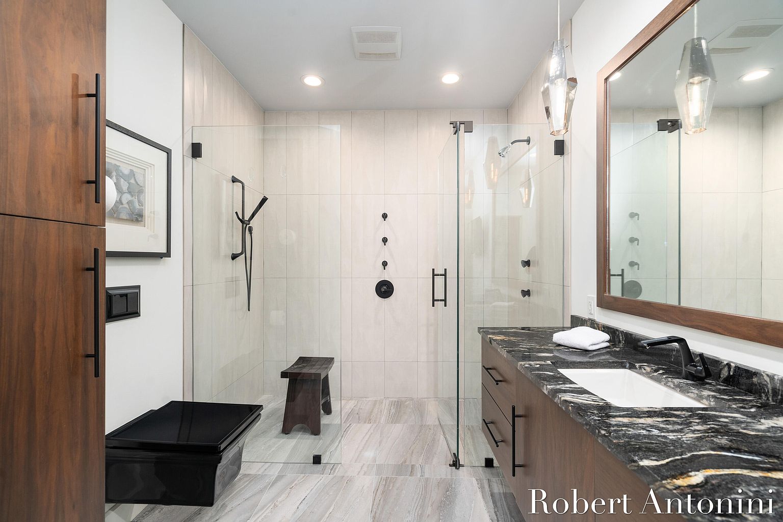 This is a modern primary bathroom featuring a glass-enclosed shower with black fixtures and a built-in bench. The vanity has a dark countertop with a white sink and black faucet, complemented by a wood-framed mirror. The flooring is light-colored tile, and the overall design is sleek and contemporary.