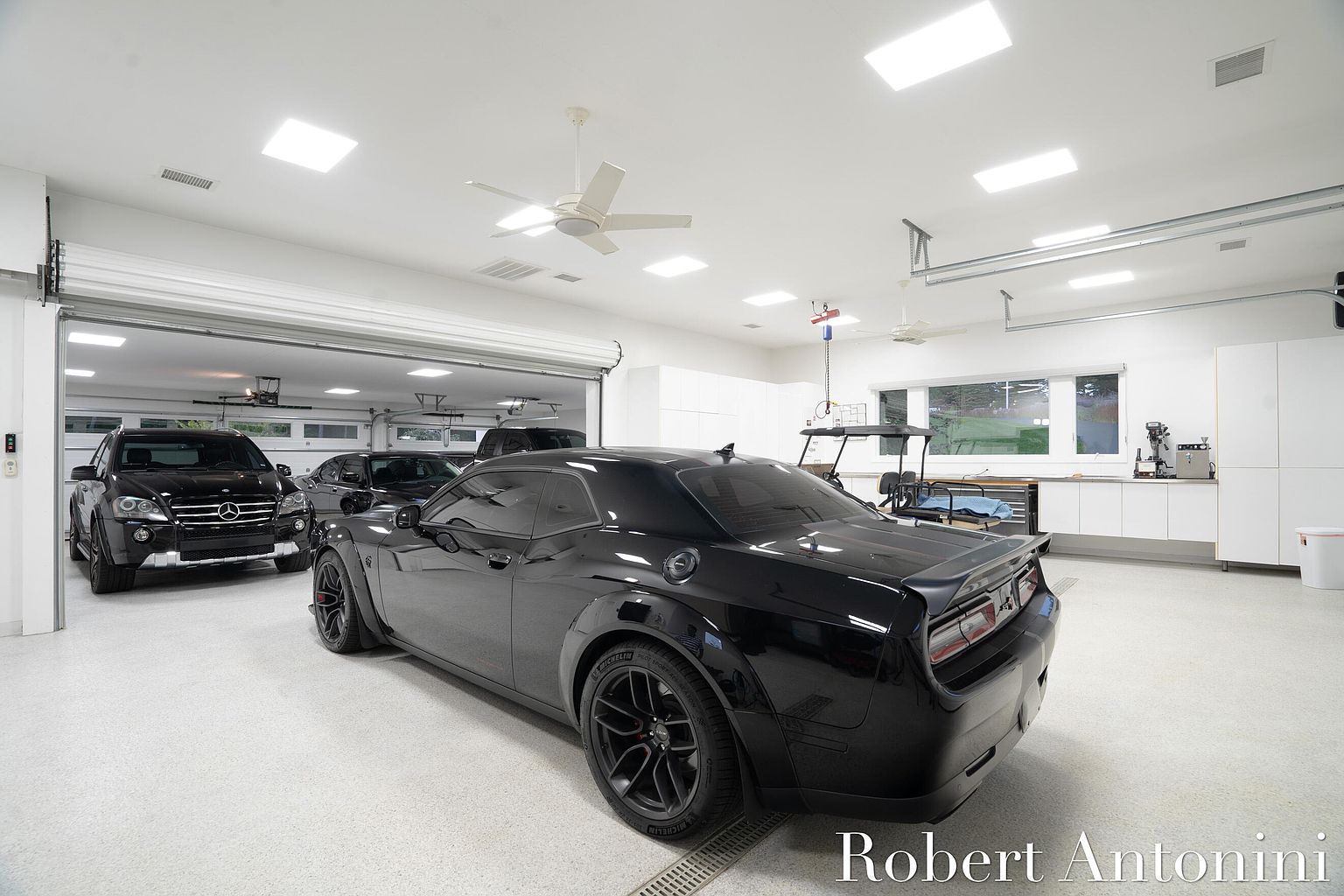 This image showcases a spacious and well-lit garage featuring multiple luxury vehicles, including a black Dodge Challenger in the foreground. The garage boasts a clean, modern aesthetic with white walls, recessed lighting, and a polished floor. The open garage door reveals additional parking space and natural light, enhancing the overall appeal of this high-end property.