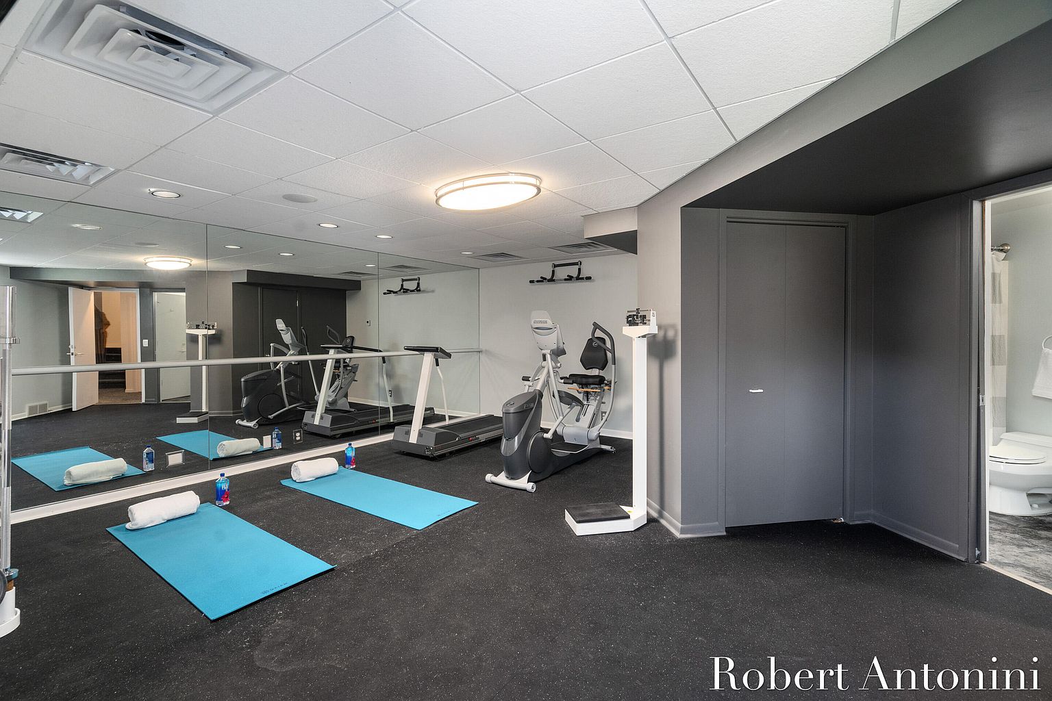 This is a well-equipped home gym featuring a treadmill, elliptical machine, and a scale. The room has a large mirror, rubber flooring, and a neutral color scheme with gray accents. A bathroom is conveniently located adjacent to the gym area, enhancing its functionality.