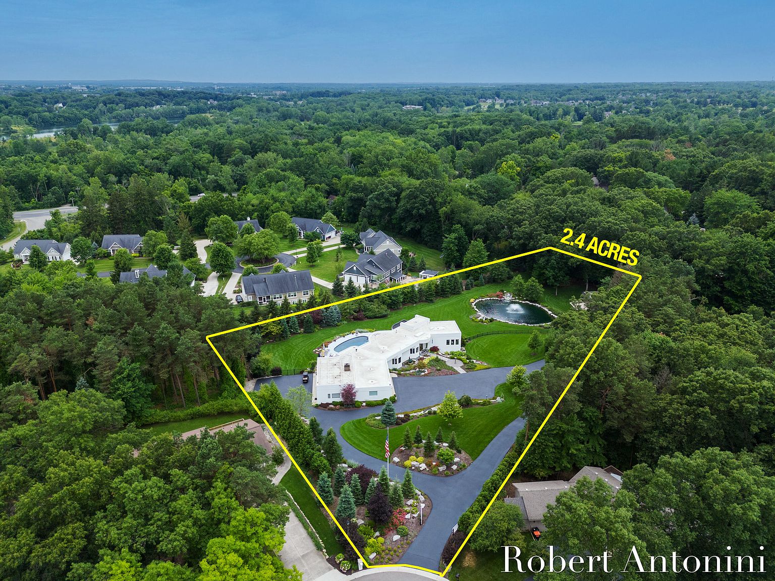 This aerial view showcases a luxurious estate situated on 2.4 acres of lush, green landscape. The property features a large, modern white house with a circular driveway, a well-manicured lawn, a pond with a fountain, and mature trees providing privacy. The overall impression is one of opulence and tranquility.