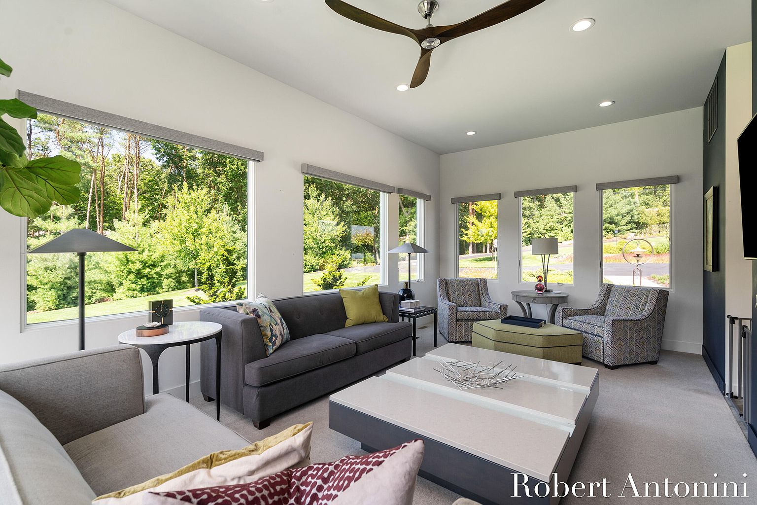 This is a bright and airy living room featuring large windows that offer views of the surrounding greenery. The room is furnished with a gray sofa, patterned armchairs, and a modern coffee table. A ceiling fan adds to the contemporary feel of the space.